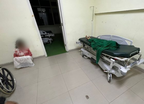 Tersangka pembunuhan terhadap anak kandung tersandar di rumah sakit dengan pakaian masih berlumuran darah, tampak mayat sang anak terbaring.